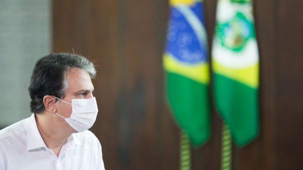 Camilo Santana está de perfil, camisa branca e usa máscara. Ao fundo, aparecem as bandeiras do Brasil e do Ceará