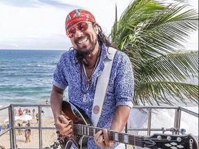 O cantor baiano de axé Bell Marques está numa praia, de bandana vermelha e camisa azul, segurando uma guitarra.