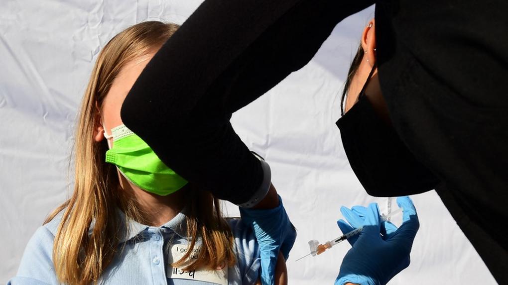 Menina loira com rosto eclipsado por braço de enfermeira sendo vacinada contra a Covid-19