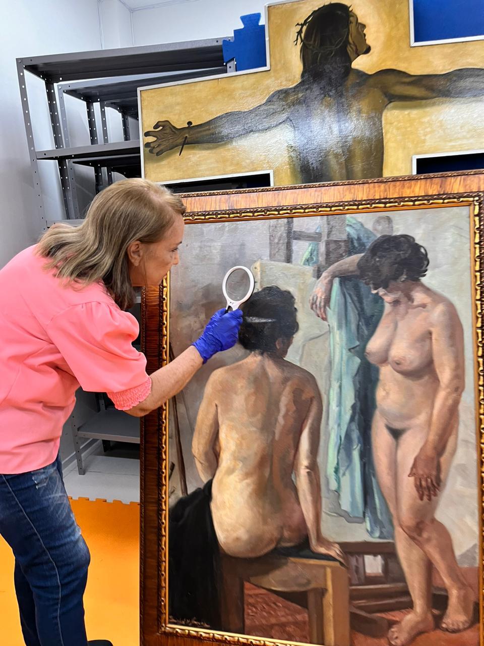 Obra “Nus no Atelier”, de Sinhá d’Amora, é analisada pela restauradora Edilma Saraiva no local em que o acervo está guardado