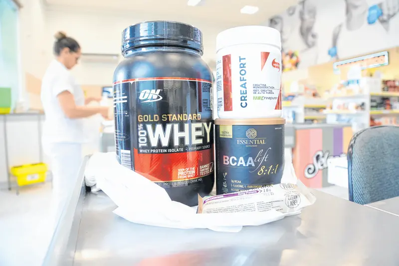 a imagem mostra um pote de whey protein e outros suplementos dentro de uma sacola e, ao fundo, um ambiente de supermercado com produtos para vida saudável
