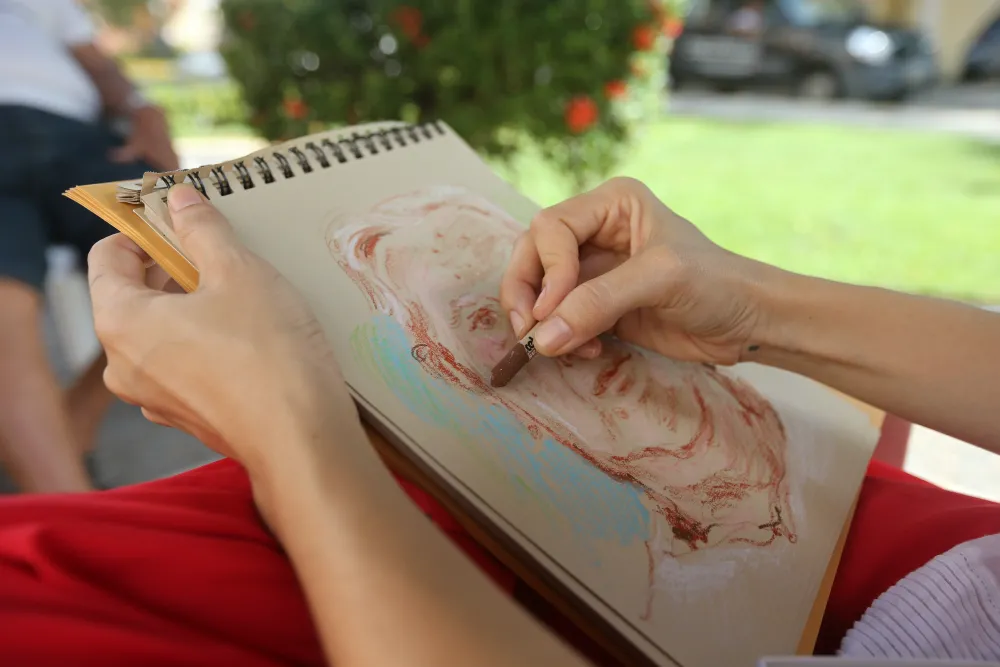 O desenho com pastel oleoso sobre papel craft traz, em nuances coloridas, as marcas do rosto de quem se dispõe a contar a própria história para Raisa Christina. A ilustradora, a partir de escuta atenta, mergulha em memórias cheias de afeto e saudade