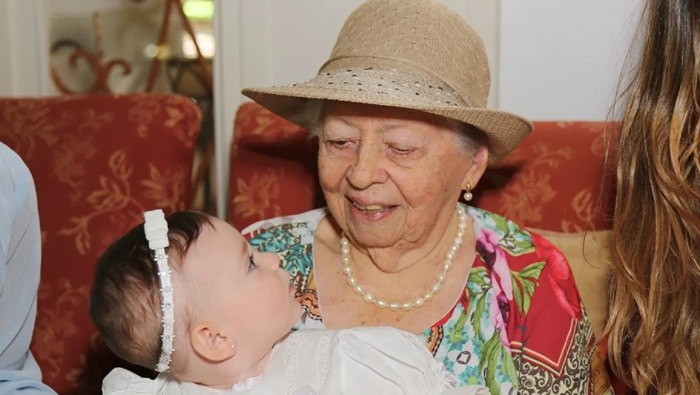 Dona Clélia Prata segura a bisneta, Lis. O vestido do batizado, objeto querido, atravessa gerações