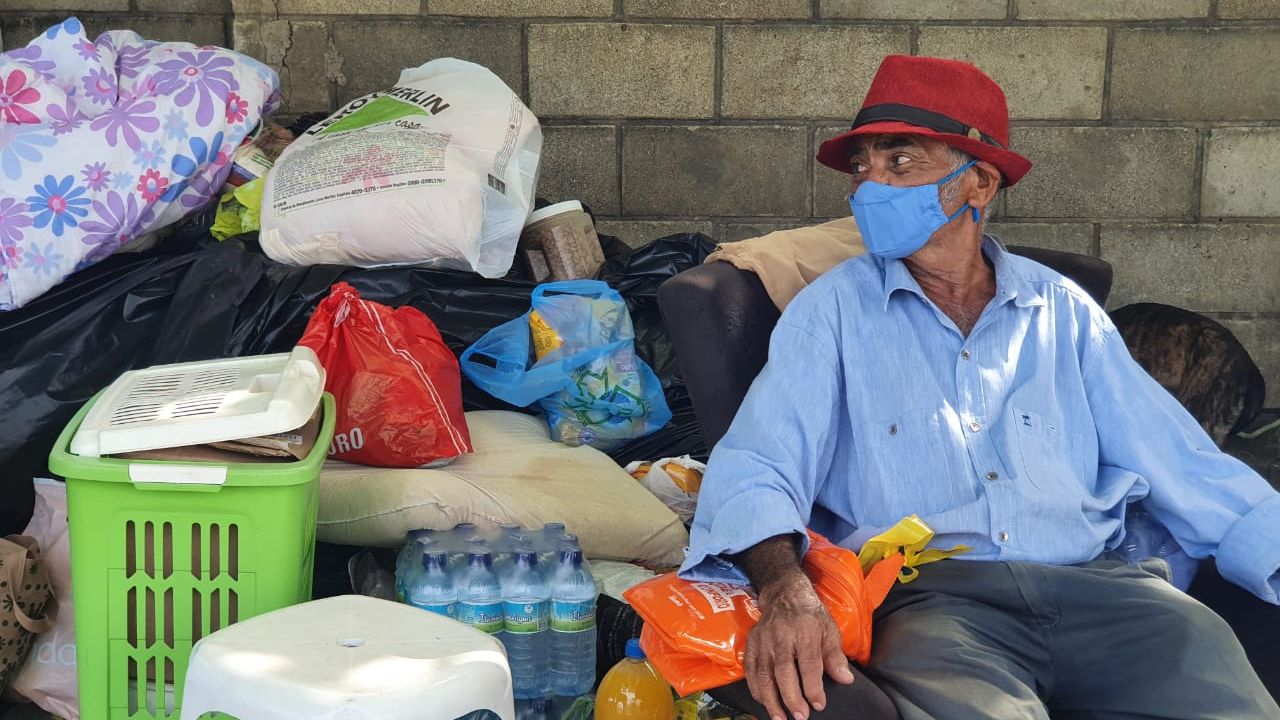 Um homem idoso de chapéu vermelho está sentado em uma calçada. Ao fundo, doações enviadas por voluntários. Entre os materiais estão garrafas de água e sacolas com roupa.