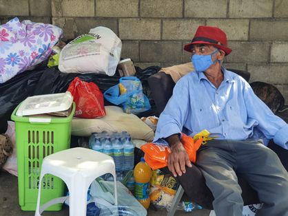 Um homem idoso de chapéu vermelho está sentado em uma calçada. Ao fundo, doações enviadas por voluntários. Entre os materiais estão garrafas de água e sacolas com roupa.