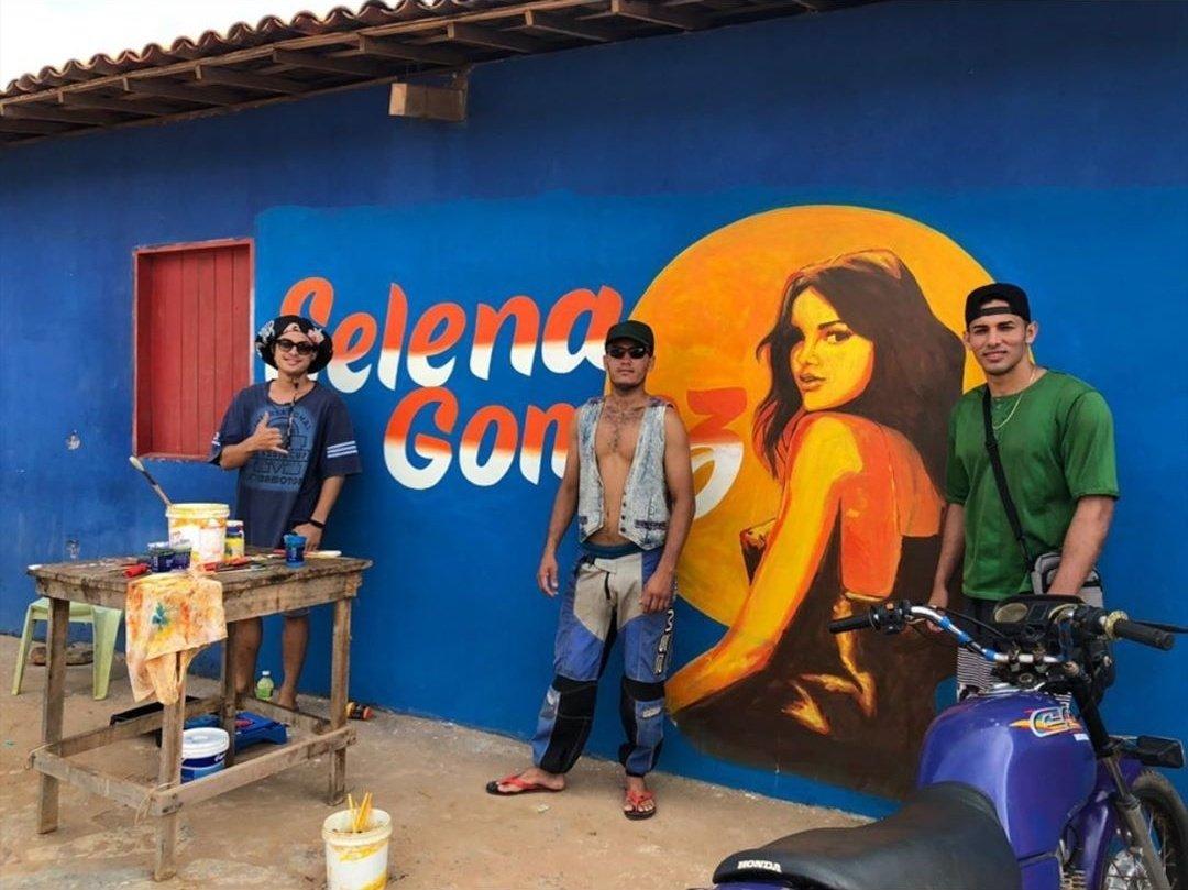 Imagem de um mural grafitado com o nome e a imagem da cantora Selena Gomez. Três homens aparecem na frente da parede, posando para a foto.