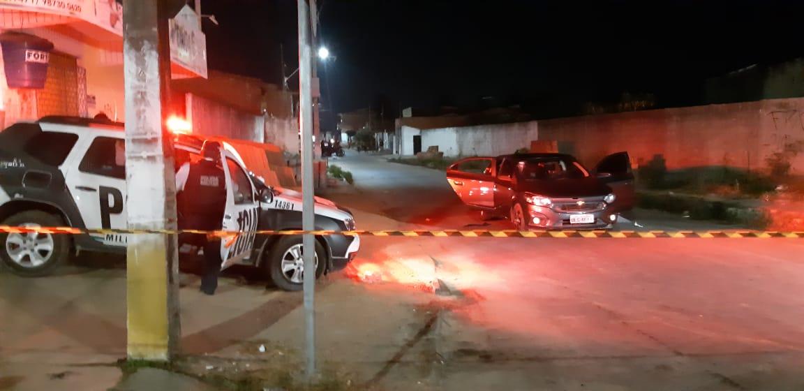 Rua vazia isolada por uma faixa policial. No lado direito há uma viatura nas cores branca e preta da polícia, com a sirene acesa, logo atrás de um poste. Do lado esquerdo da imagem há um carro preto com as portas abertas.