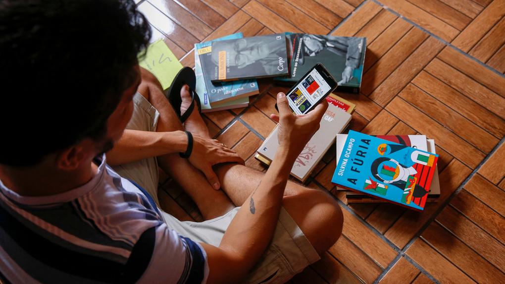 Esta é uma imagem de um leitor com um celular e alguns livros sobre o chão.
