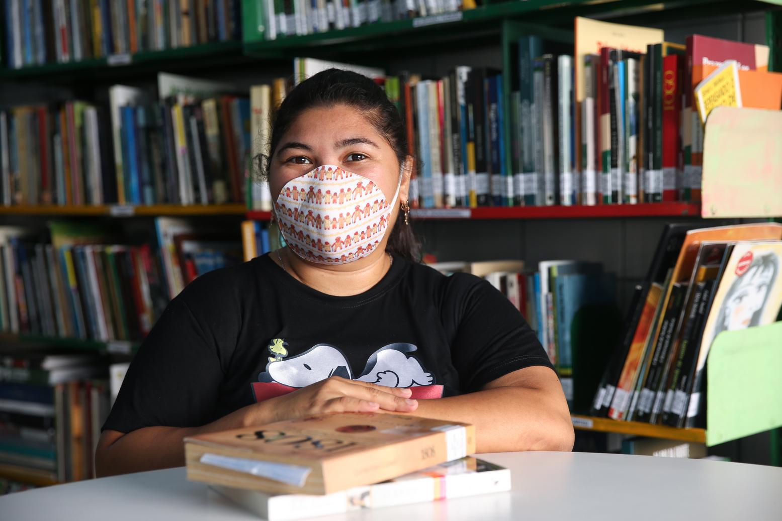 Esta é uma mulher jovem, de máscara, com alguns livros na Biblioteca Sorriso da Criança, no bairro Presidente Kennedy