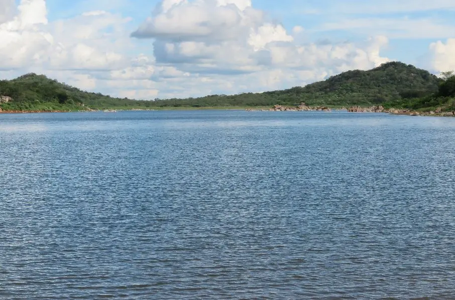 Grande açude com água azul límpida, margeada por dois montes de verdes e um céu azul com nuvens brancas