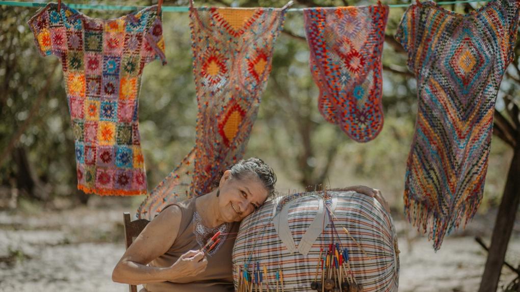 Esta é uma foto da Mestra Raimundinha. Ela está sentada com a cabeça reclinada sobre uma almofada de renda de bilro. Atrás dela, estão algumas peças de renda expostas num varal