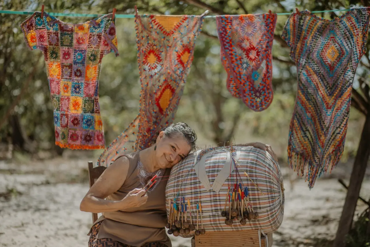 Esta é uma foto da Mestra Raimundinha. Ela está sentada com a cabeça reclinada sobre uma almofada de renda de bilro. Atrás dela, estão algumas peças de renda expostas num varal