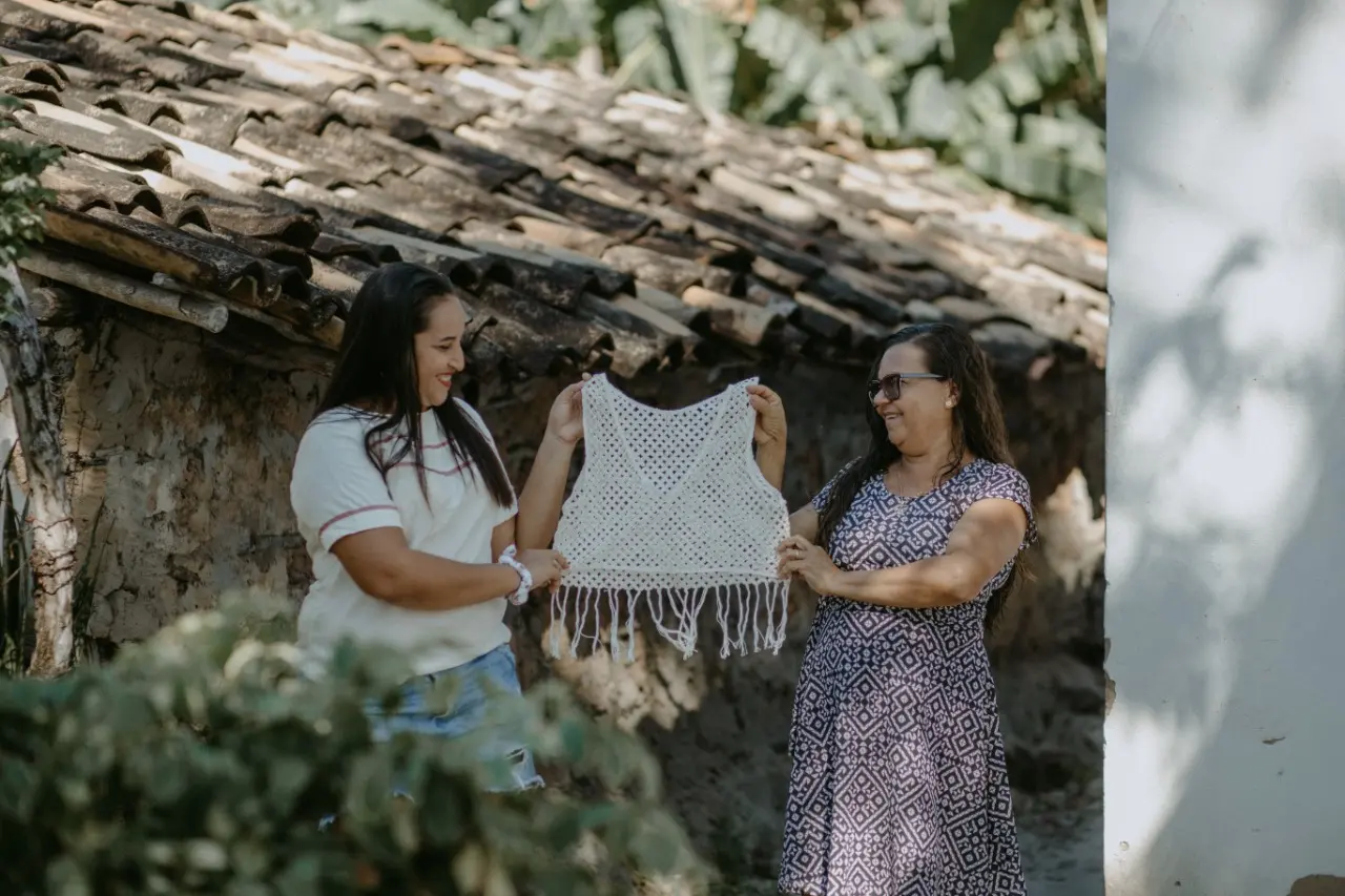 Esta é a imagem de duas rendeiras, Ivone e Terezinha. Elas estão segurando um cropped branco feito em renda de birlo
