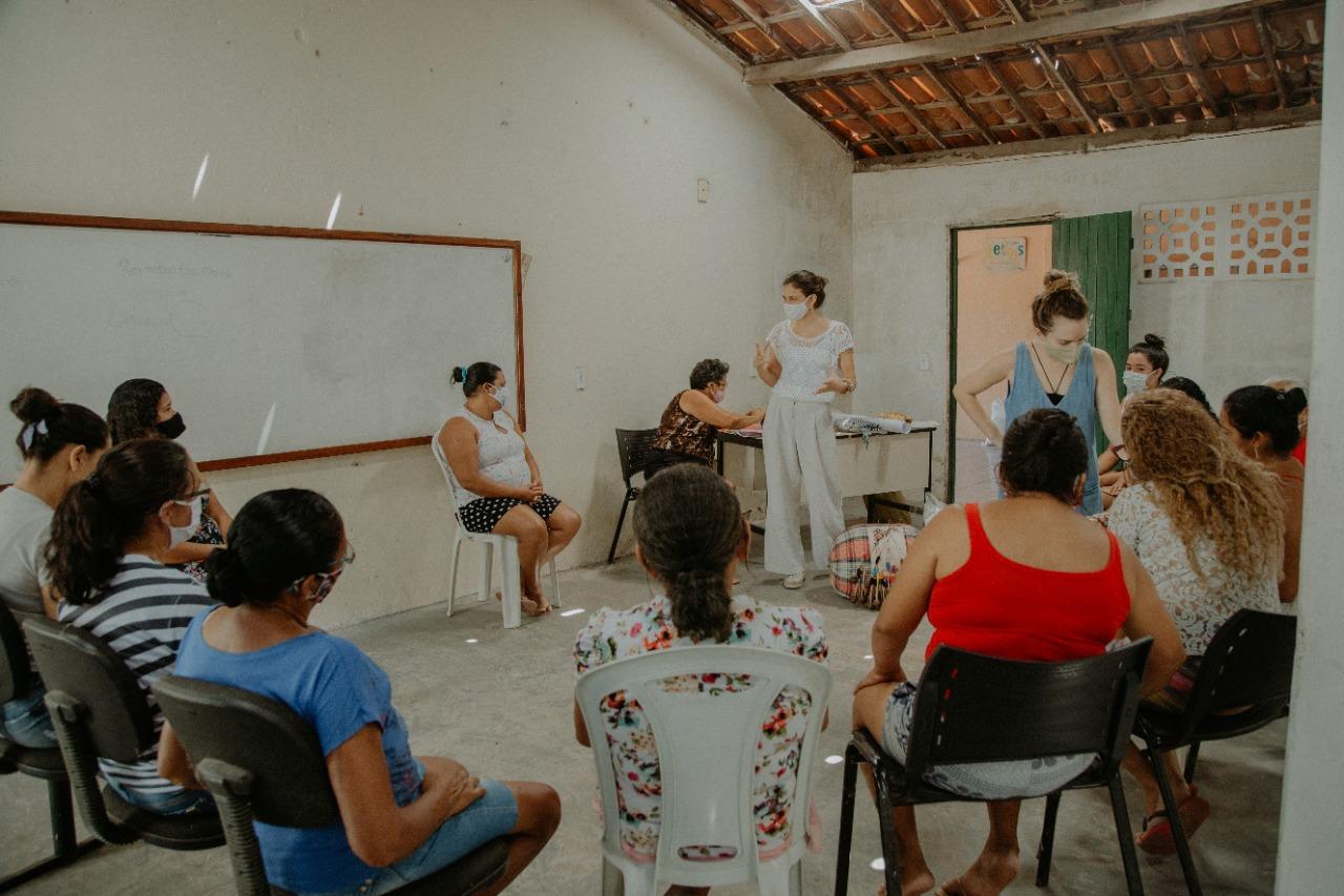 Esta é a imagem de uma oficina. Uma mulher coordena a oficina de máscara. Sentadas, também de máscara, várias artesãs acompanham a aula