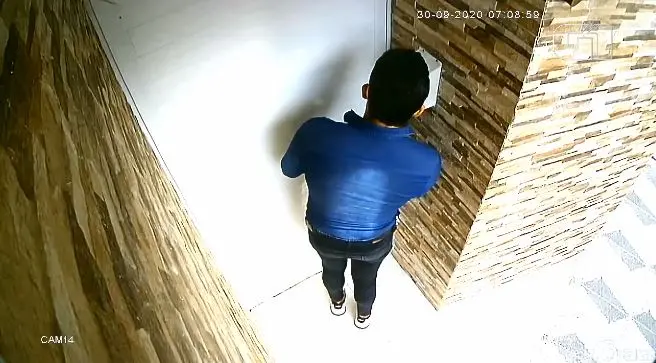 Homem vestido com camisa jeans, calça preta, tênis branco, usando óculos escuros e máscara no rosto, segurando um celular na mão, em frente à porta de um condomínio.