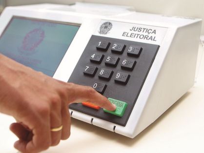 Imagem de uma urna eletrônica eleitoral, com uma mão apertando o botão verde de Confirma.