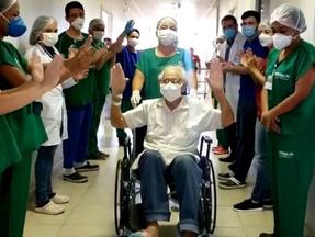 Idoso é levado em cadeira de rodas, com os braços levantados, comemorando, enquanto vários enfermeiros estão ao redor aplaudindo.