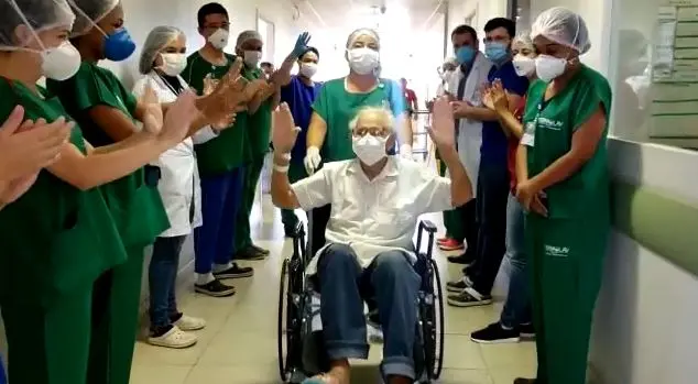 Idoso é levado em cadeira de rodas, com os braços levantados, comemorando, enquanto vários enfermeiros estão ao redor aplaudindo.