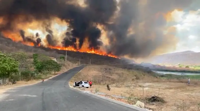 Em primeiro plano, uma estrada que corta uma área de vegetação rasteira e segue rumo a um monte com chamas altas. Um carro e algumas pessoas estão parados na rodovia, observando o incêndio.