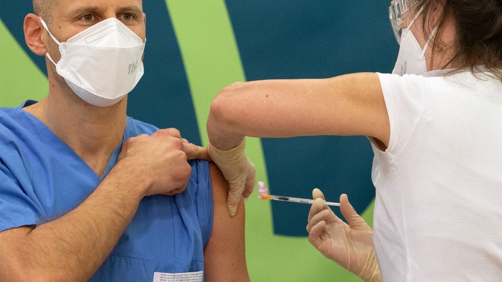 Homem de bata azul, possivelmente profissional de saúde, com uma máscara branca de proteção cobrindo o nariz e a boca, recebe a vacina contra a Covid-19 no braço esquerdo. A vacina é aplicada por uma mulher com uma camisa branca, também de máscara de proteção.