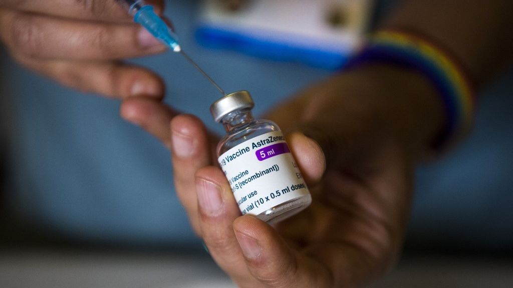 Imagem mostra as mãos de uma pessoa segurando uma pequena ampola da vacina contra a Covid-19, com uma seringa sendo injetada para colher uma dose da vacina.