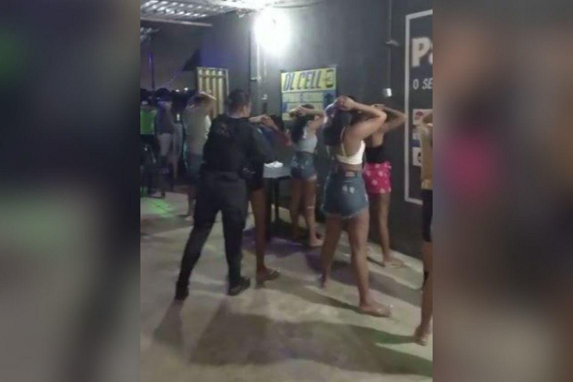 Várias pessoas, a maioria mulheres, de costas, com as mãos na cabeça e as pernas entreabertas, sendo revistadas por uma policial