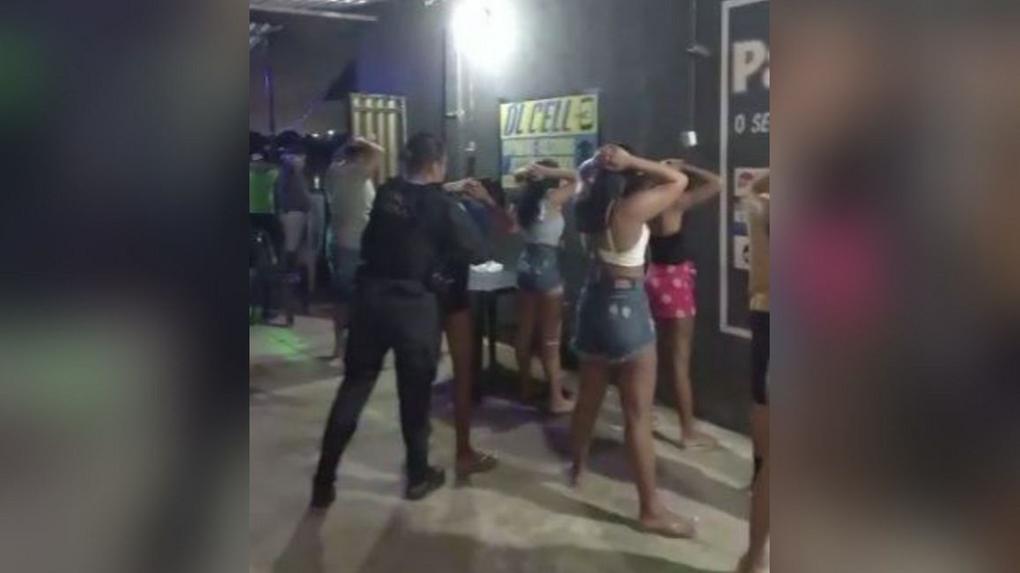 Várias pessoas, a maioria mulheres, de costas, com as mãos na cabeça e as pernas entreabertas, sendo revistadas por uma policial