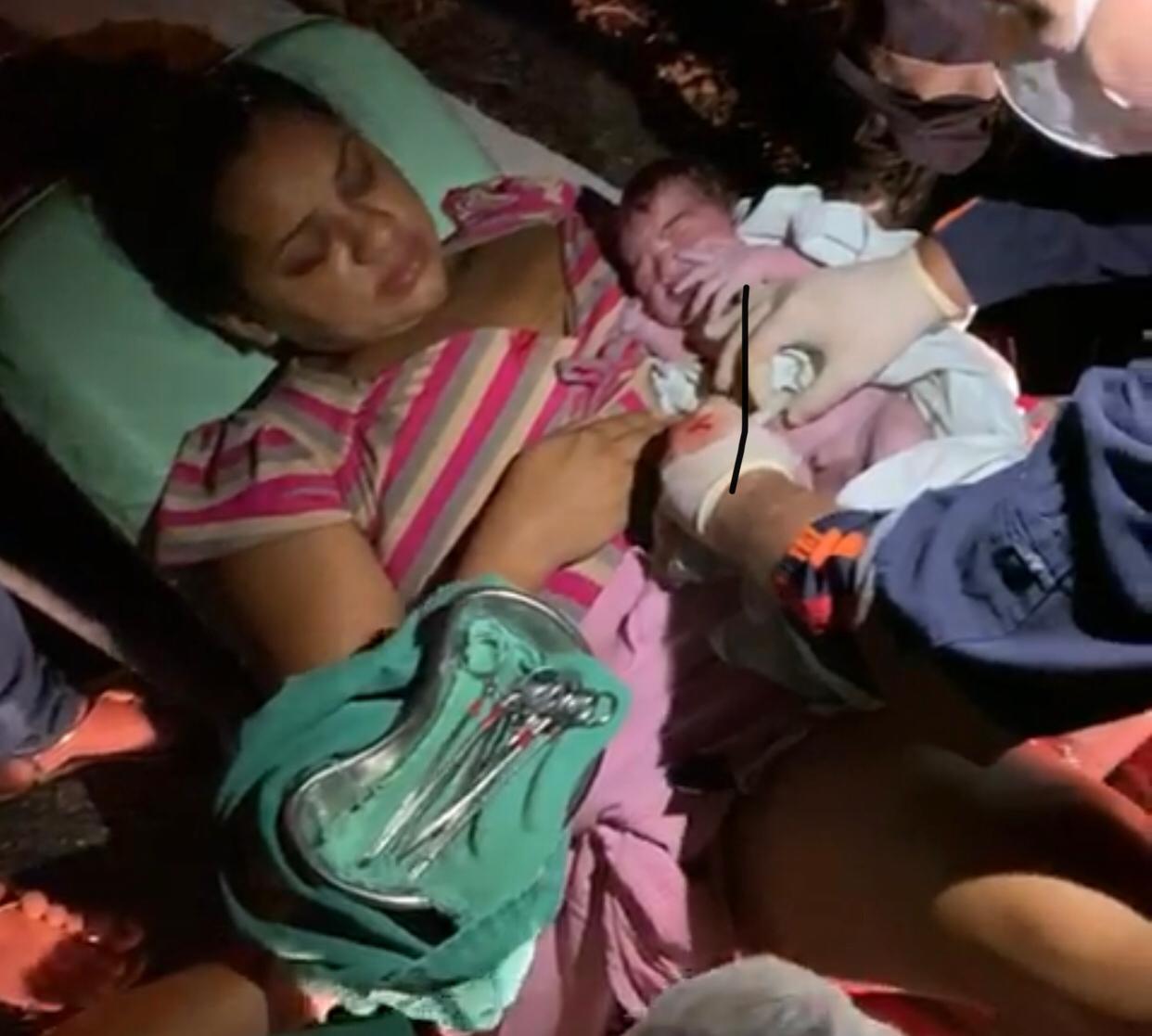 Mulher que acabou de ter um bebê segura a criança nos braços, ainda deitada na maca, enquanto a criança é examinada por médicos