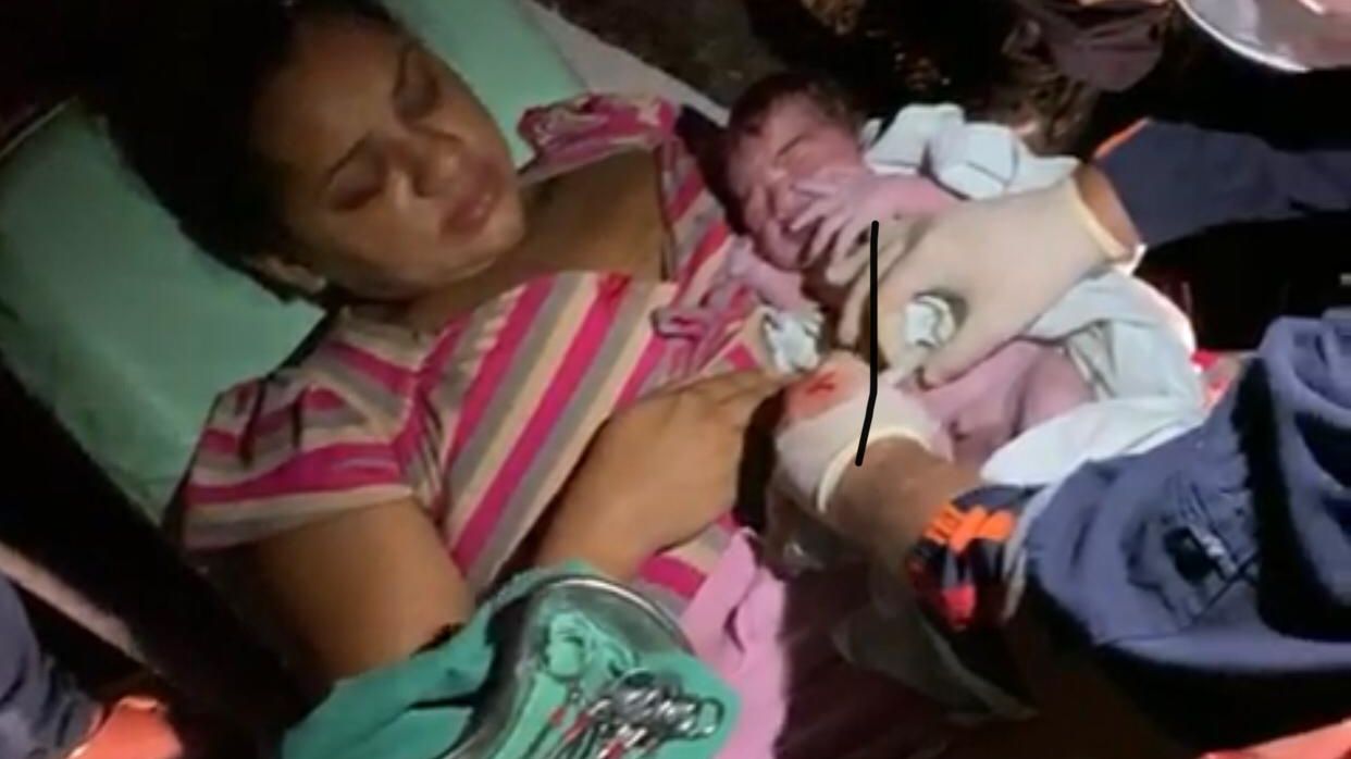 Mulher que acabou de ter um bebê segura a criança nos braços, ainda deitada na maca, enquanto a criança é examinada por médicos