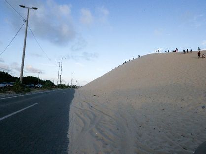 Grande duna de areia margeando uma rodovia. Algumas pessoas estão em cima da duna.