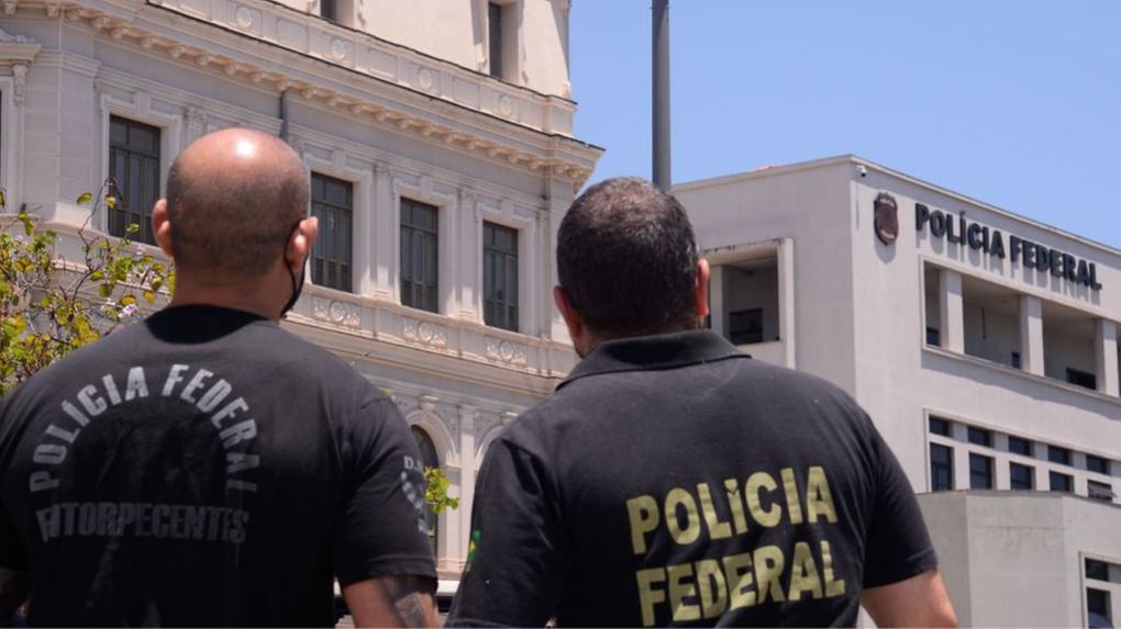 No primeiro plano da imagem, há dois agentes da Polícia Federal de costas. Em segundo plano, está o prédio da Polícia Federal.