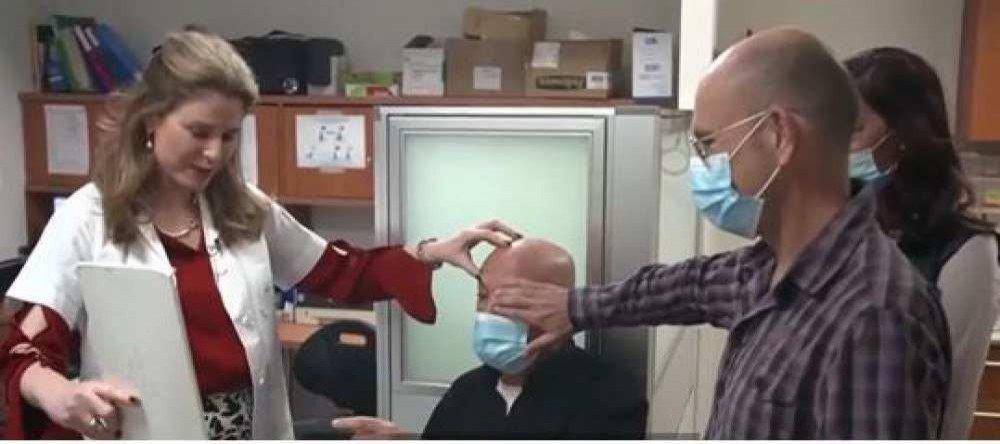 Está imagem mostra três pessoas. Da esquerda para a direita: uma médica, um idoso de máscara cirúrgica e um homem de máscara cirúrgica. O idoso lê um papel após recuperar a visão ao receber o primeiro transplante de córnea artificial do mundo.