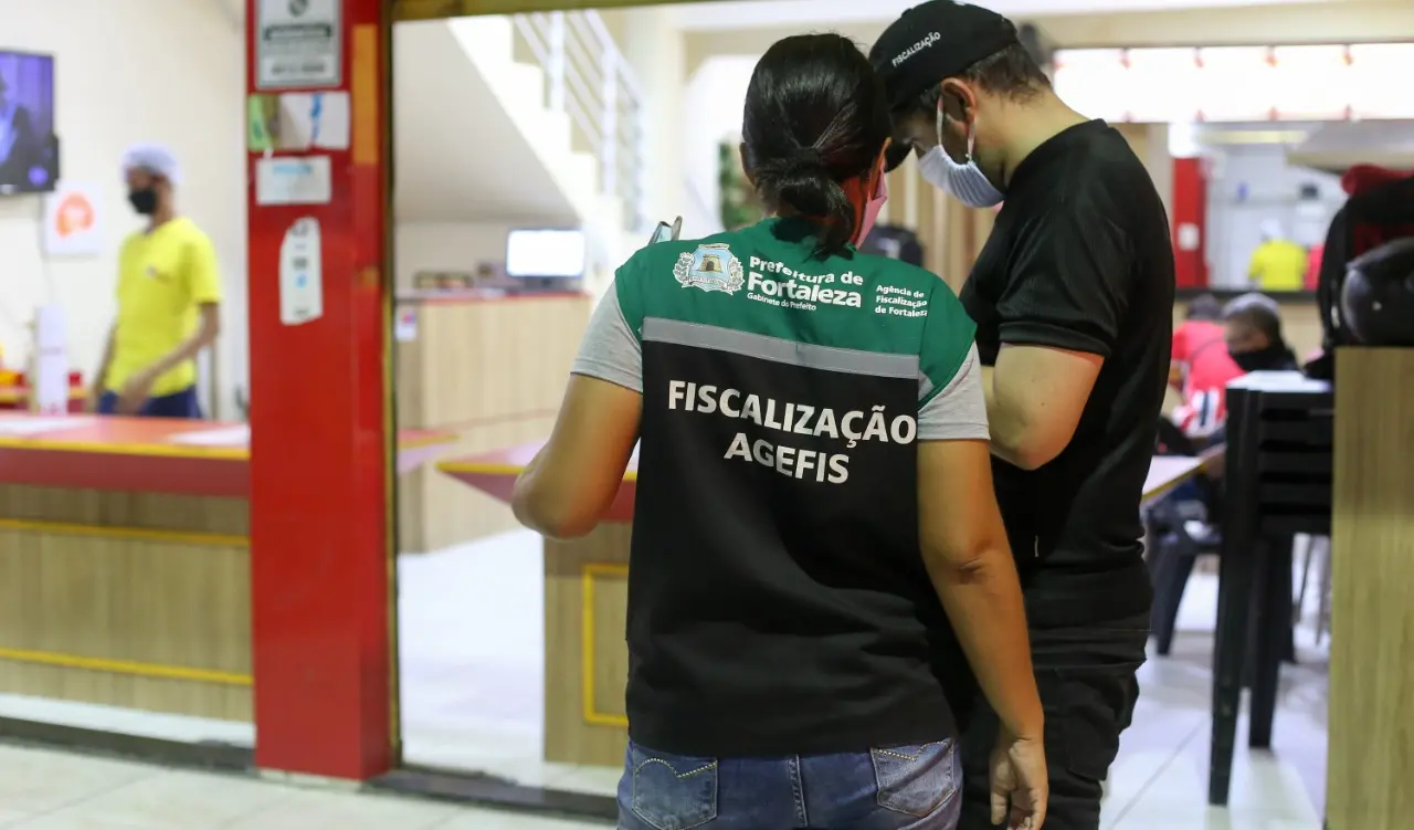 Nesta imagem há, em primeiro plano, uma mulher e um homem de costas, ambos de máscara cirúrgica. A mulher está vestindo um colete que a identifica como servidora da Agência de Fiscalização de Fortaleza e o homem está de camisa preta. No segundo plano, há uma restaurante.