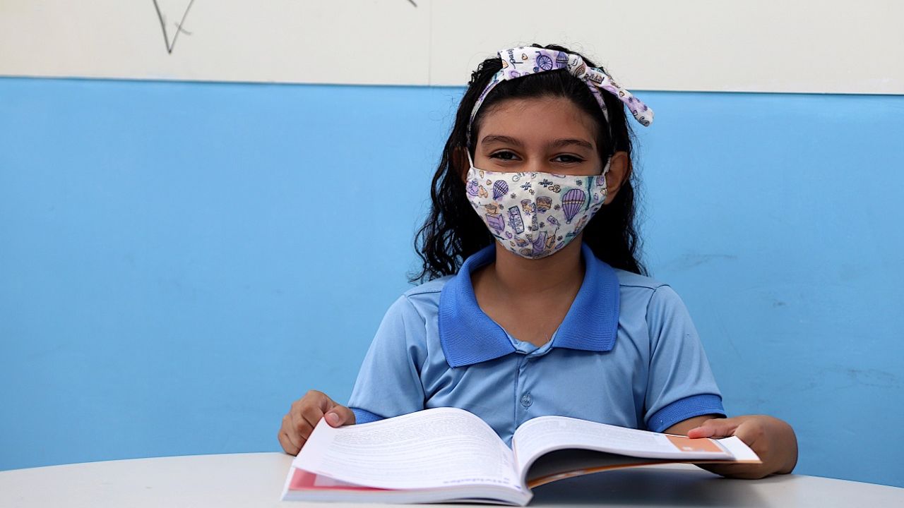 Esta é a imagem de uma menina vestida com o fardamento das escolas públicas da Prefeitura de Fortaleza. Ela está sentada de frente a uma mesa segurando um livro  e usando máscara.