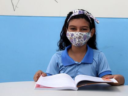 Esta é a imagem de uma menina vestida com o fardamento das escolas públicas da Prefeitura de Fortaleza. Ela está sentada de frente a uma mesa segurando um livro  e usando máscara.