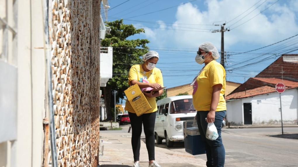 Esta é a imagem de duas profissionais de saúde vestindo blusas amarelas e usando equipamentos de proteção, como máscaras e toucas, visitam domicílios em Fortaleza durante campanha de vacinação contra a Covid-19.