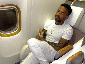 esta imagem mostra o cantor Nego do Borel, usando blusa e calça branca, sentando em uma cadeira luxuosa cadeira de uma aeronave. Ele está segurando uma taça com bebida e está com uma expressão neutra.