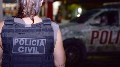 Policial Civil mulher de costas usando colete à prova de balas com o nome da Polícia Civil. Ao fundo, viatura da Polícia Civil estacionada