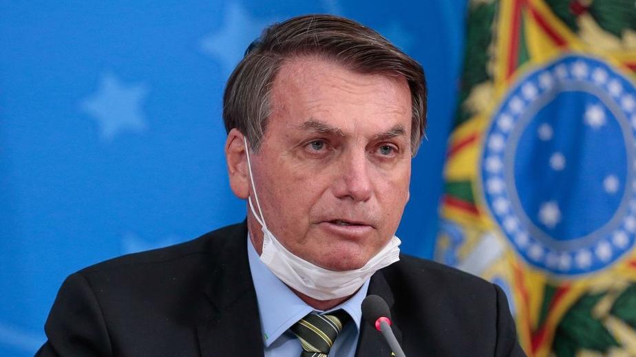 Bolsonaro está com uma máscara no queixo, olhar distante e boca semiaberta. No fundo, aparece a bandeira do Brasil