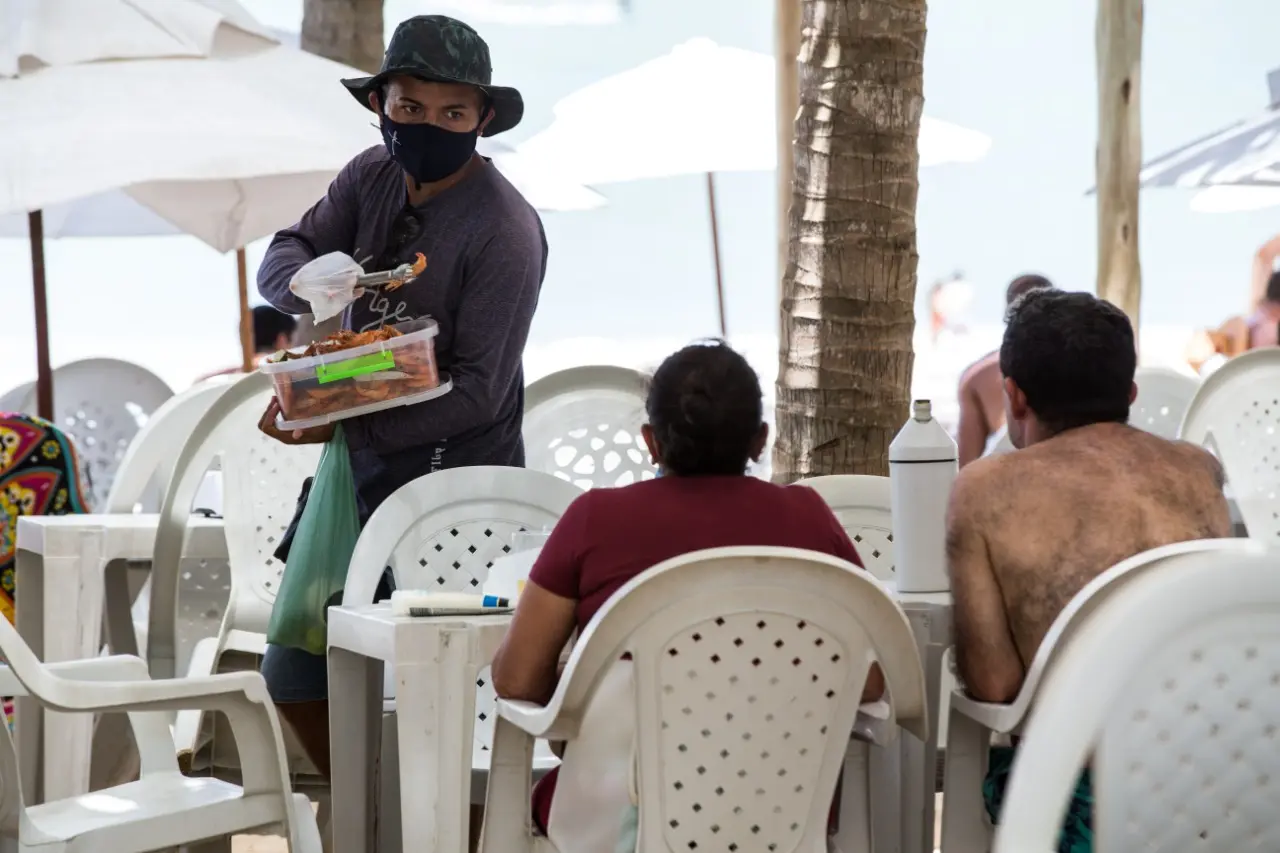 Na praia, um vendedor ambulante oferece camarão a um casal que está sentado em cadeiras de uma barraca de praia de frente para o mar