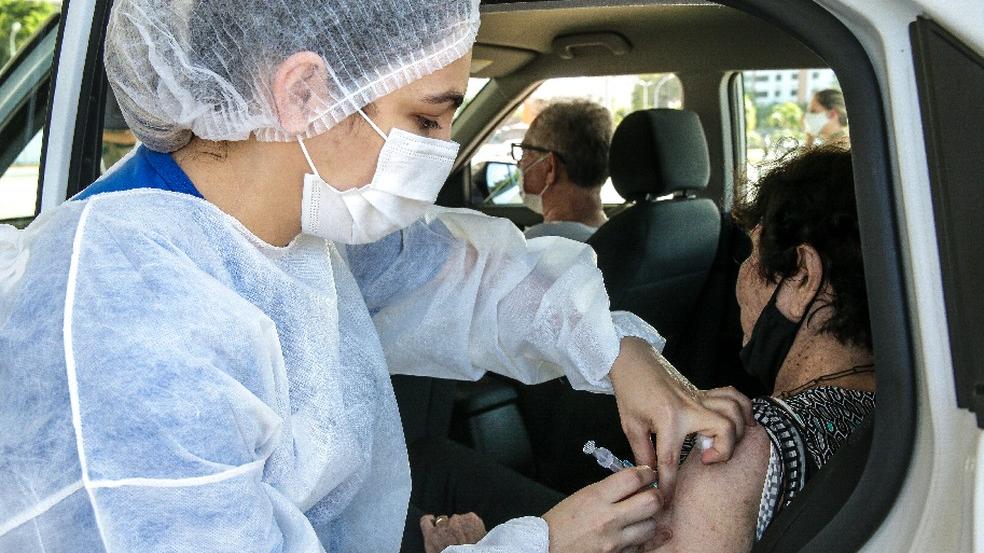 Profissional de saúde usando touca, máscara e bata cirúrgicas aplicando vacina contra Covid-19 em uma idosa em um drive-thru