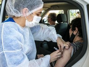 Profissional de saúde usando touca, máscara e bata cirúrgicas aplicando vacina contra Covid-19 em uma idosa em um drive-thru