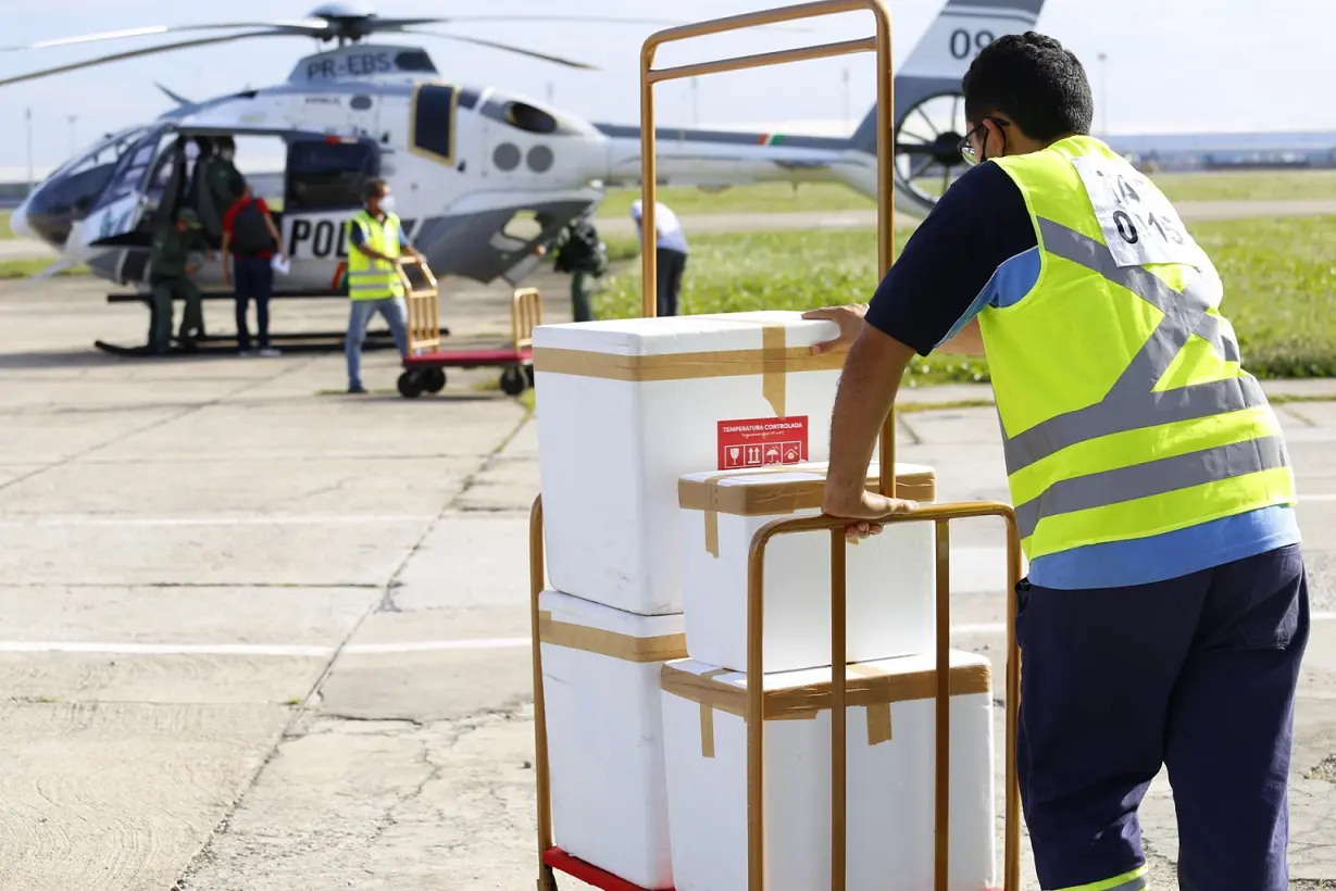 Homem empurra carrinho com quatro isopores brancos com vacinas. Ele segue em direção a um helicóptero da Polícia Militar que leva os imunizantes para cidades do interior