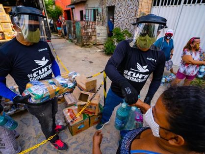Homens de máscara e face shield entregam cestas básicas a uma mulher de máscara na favela