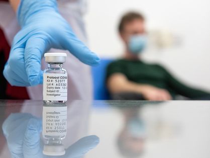 Frasco com o líquido da vacina contra a Covid-19 preste a ser pegue por uma mão com luva azul, à frente de um paciente