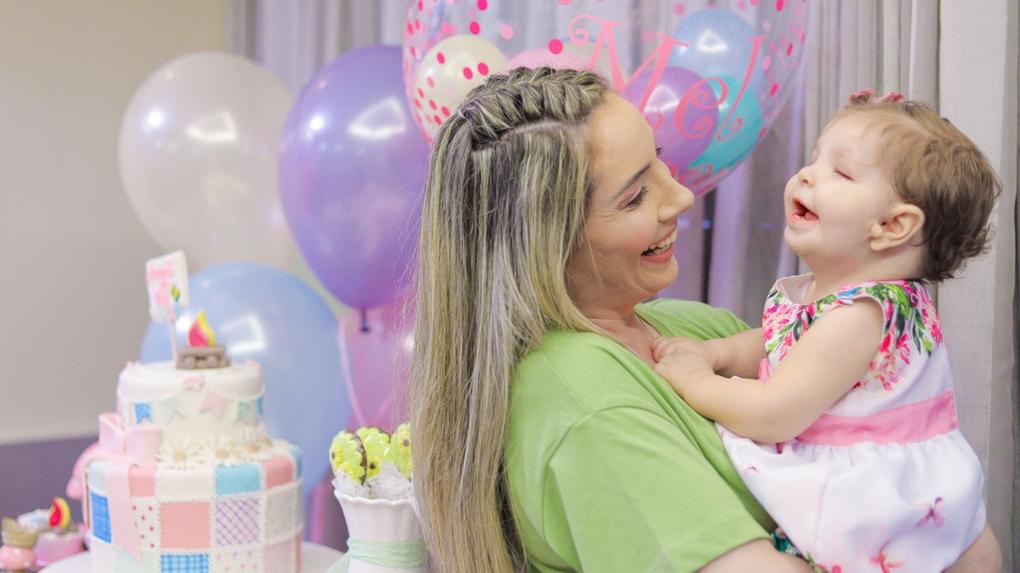 Marcela sorri enquanto segura a filha Melina nos braços na frente do bolo de aniversário da bebê