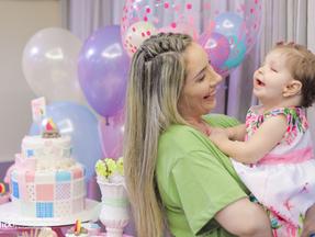 Marcela sorri enquanto segura a filha Melina nos braços na frente do bolo de aniversário da bebê