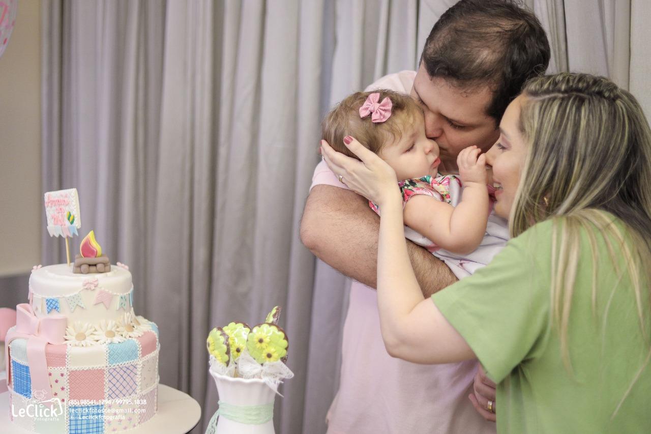 Fabrício, marido de Marcela, segura e beija a cabeça de Melina enquanto a mãe conversa com a bebê. A família aparece ao lado de um bolo de aniversário e balões decorativos