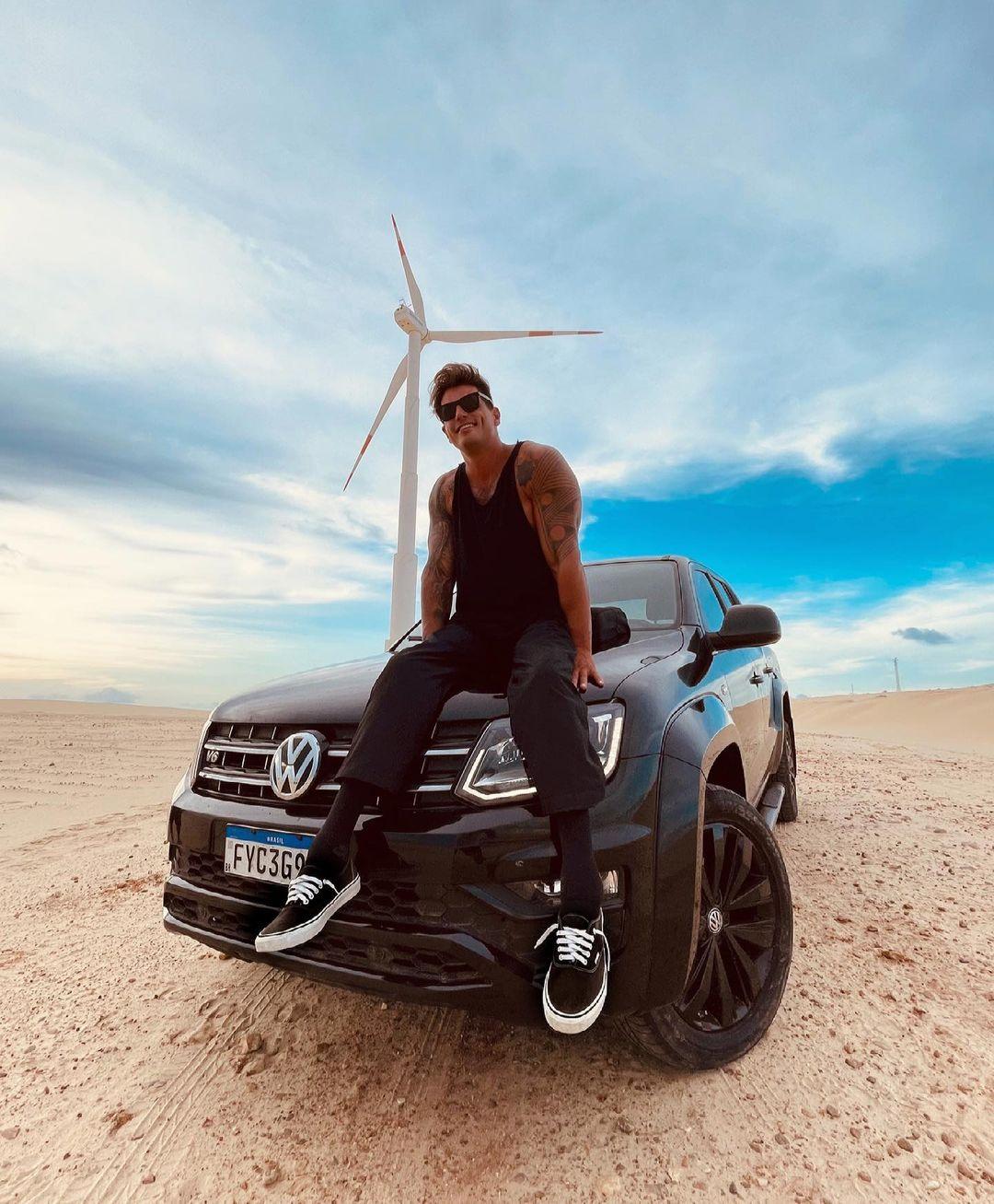 Fernando Fernandes sentado na frente do carro com uma turbina de energia eólica ao fundo localizada em Taíba