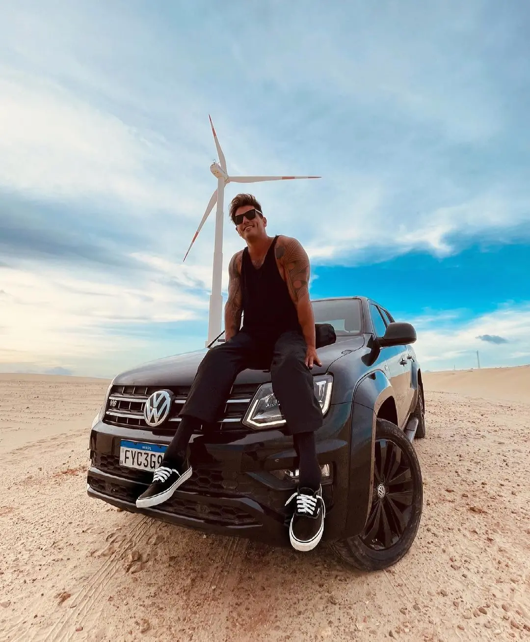 Fernando Fernandes sentado na frente do carro com uma turbina de energia eólica ao fundo localizada em Taíba